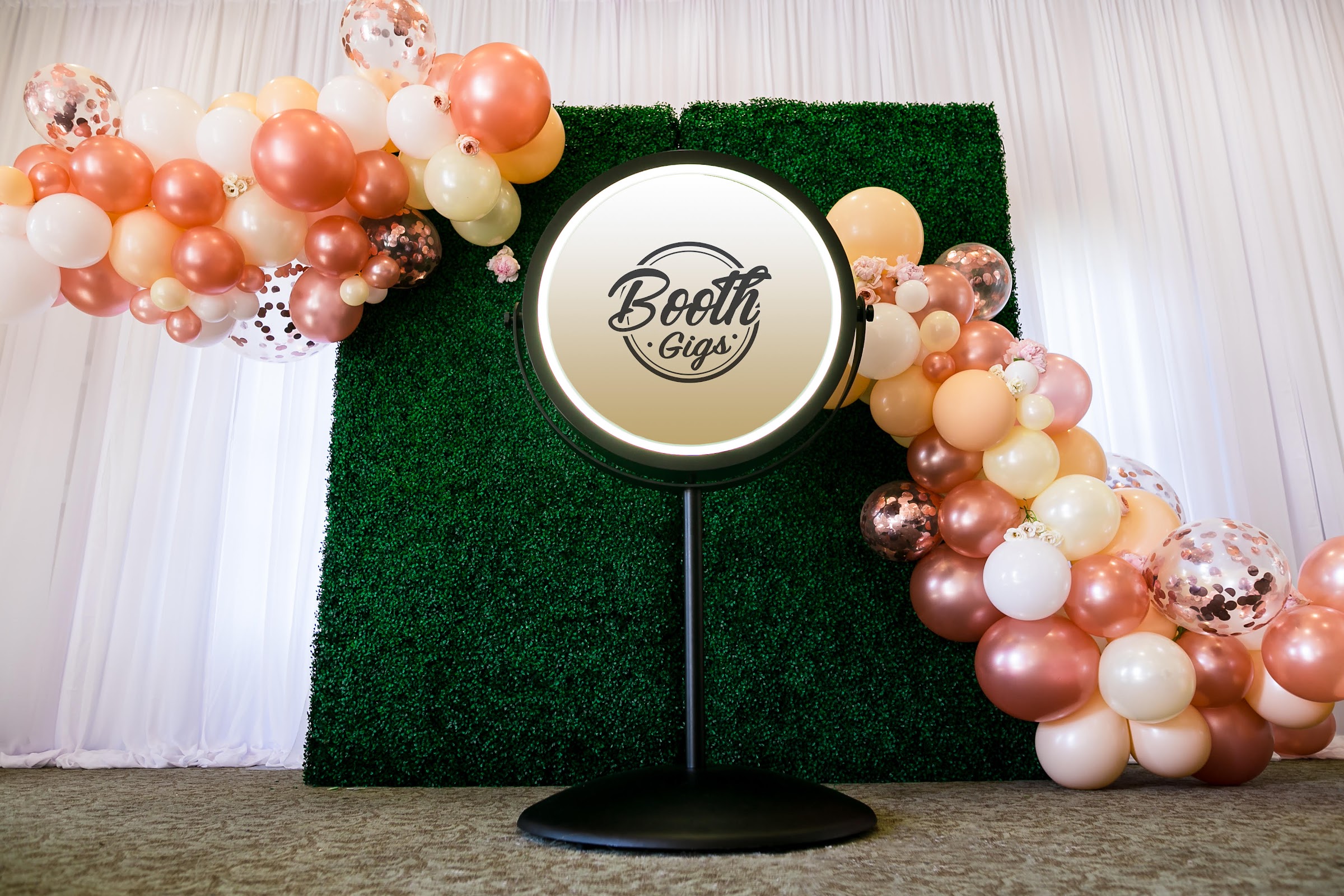 Mirror booth showing the Booth Gigs logo in front of a greenery wall and rose-gold balloon garland