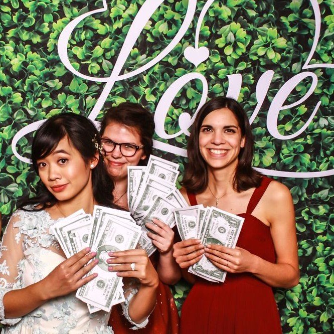 Three guests fanning prop money in front of a Love greenery wall
